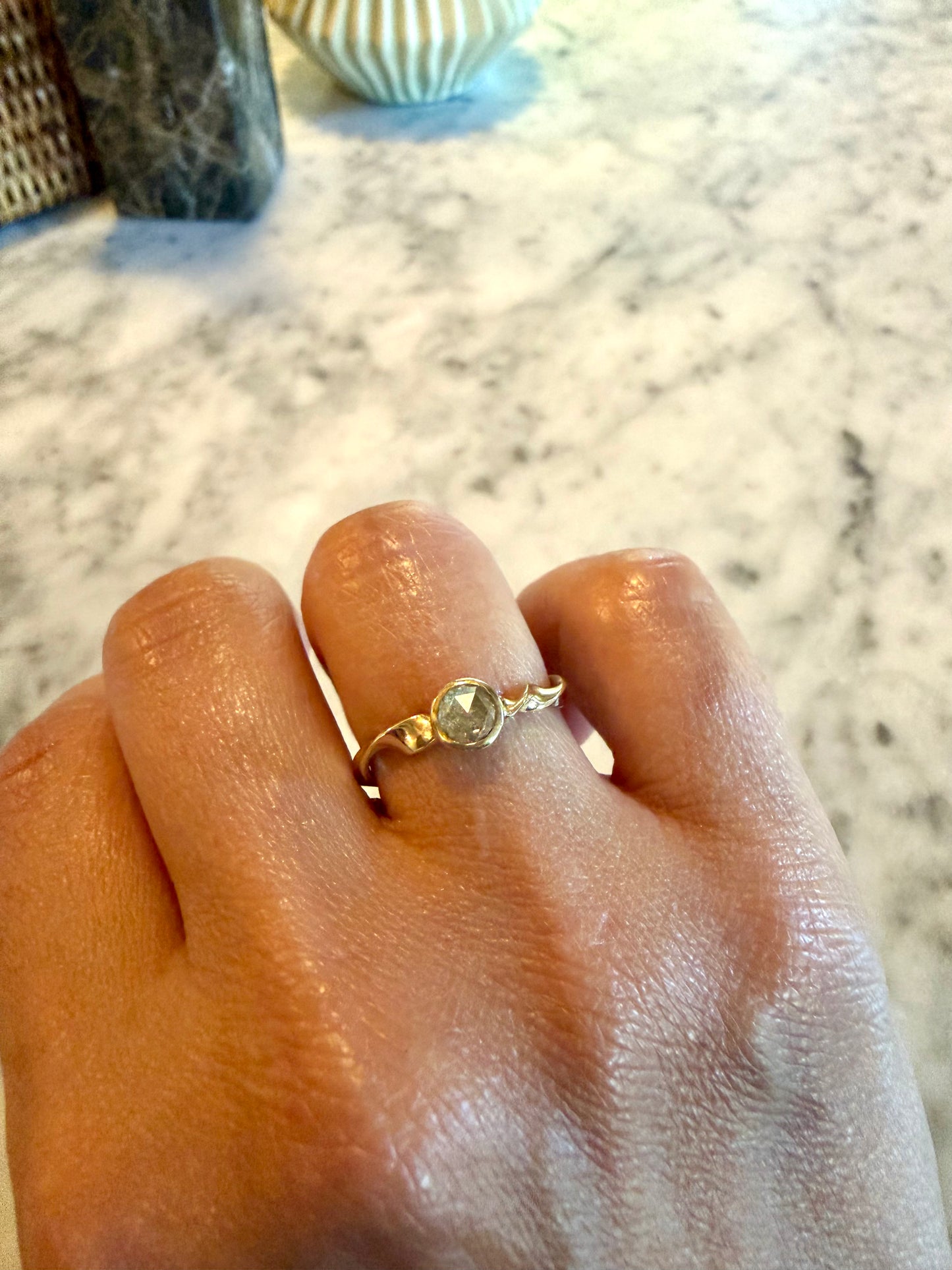 A gold ring with a rosecut gray diamond worn on a person's finger, shown against a marble background.