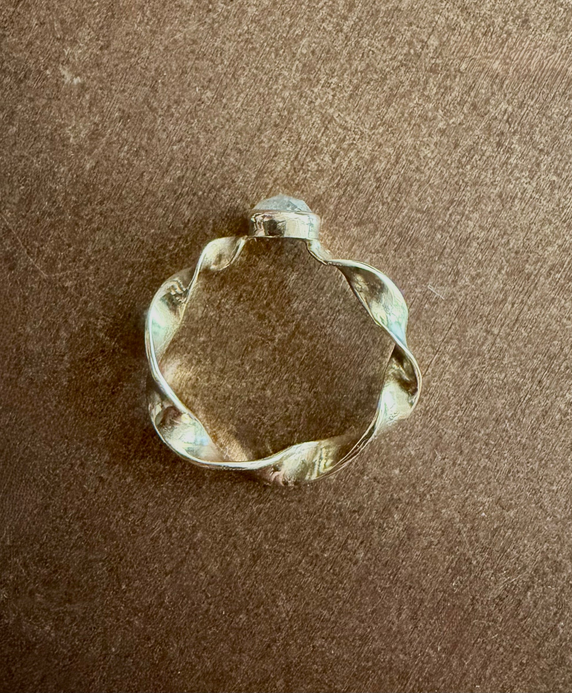 A gold ring with a twisted, organic shape, and a gray diamond photographed on a brown textured surface.