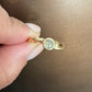 A close-up of a hand holding a small gold ring with a round gray diamond against a brown background.