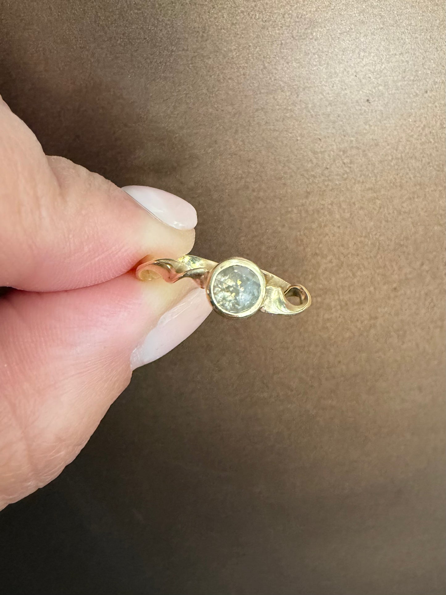 A close-up of a hand holding a small gold ring with a round gray diamond against a brown background.