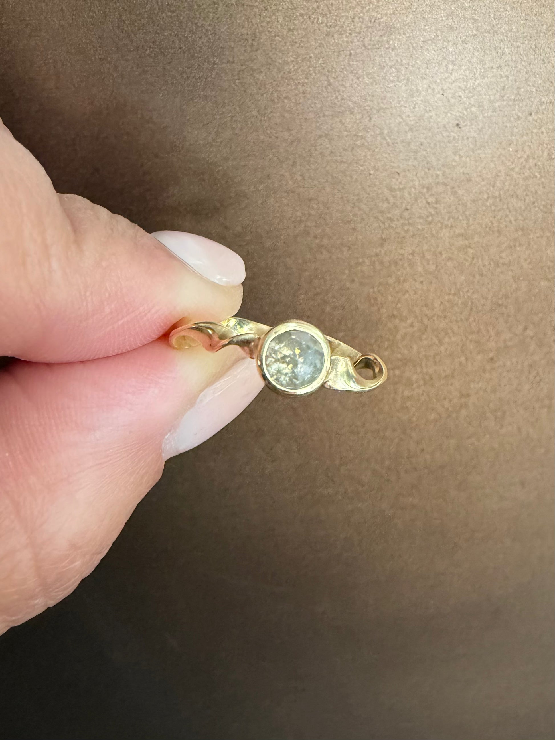 A close-up of a hand holding a small gold ring with a round gray diamond against a brown background.