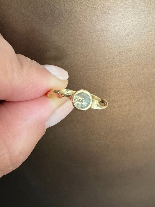 A close-up of a hand holding a small gold ring with a round gray diamond against a brown background.